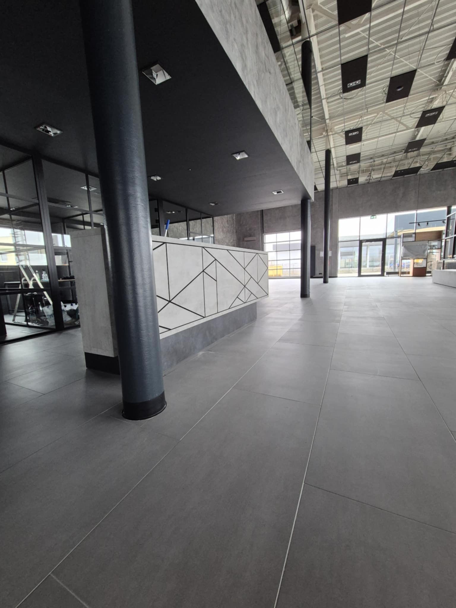 Przestronne wnętrze biurowe z szarą podłogą, czarnymi kolumnami i recepcją z geometrycznym wzorem, widok na przeszklone biura i wejście.
