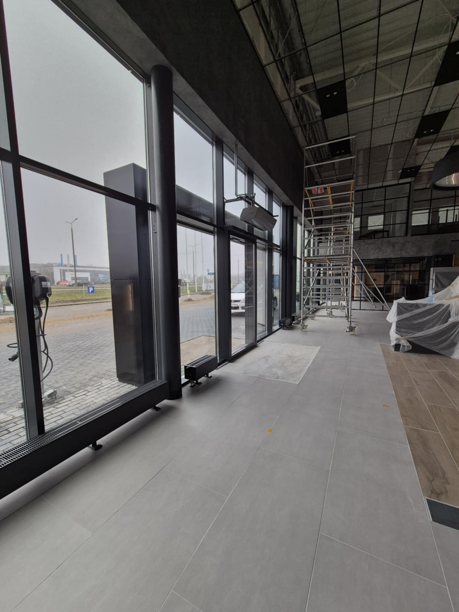 Wnętrze biura w trakcie wykańczania, widoczne duże okna z widokiem na parking, metalowe rusztowanie i zakryte folią meble.