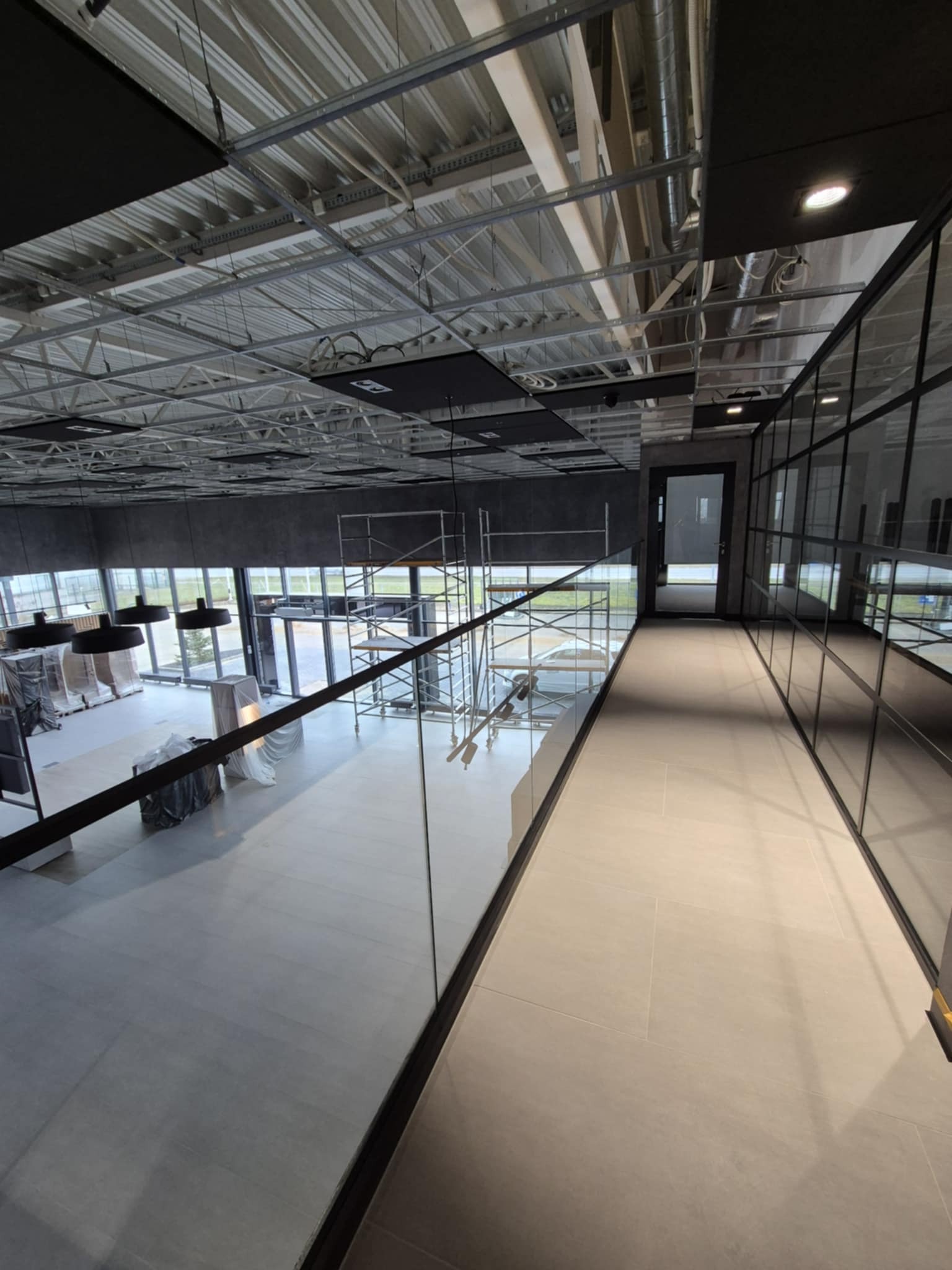 Wnętrze biurowe w trakcie wykańczania; widoczne rusztowania, szklane ścianki działowe, sufit z instalacjami i lampy wiszące.