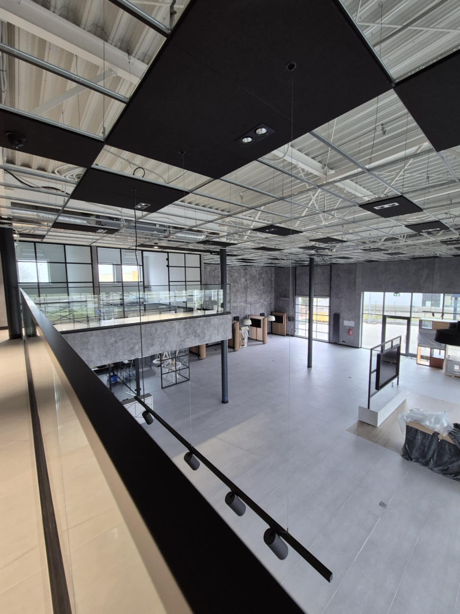 Wnętrze biurowe po remoncie, widok z antresoli na otwartą przestrzeń z szarą podłogą i sufit z czarno-białymi panelami akustycznymi, widoczne elementy konstrukcyjne i instalacje pod sufitem.