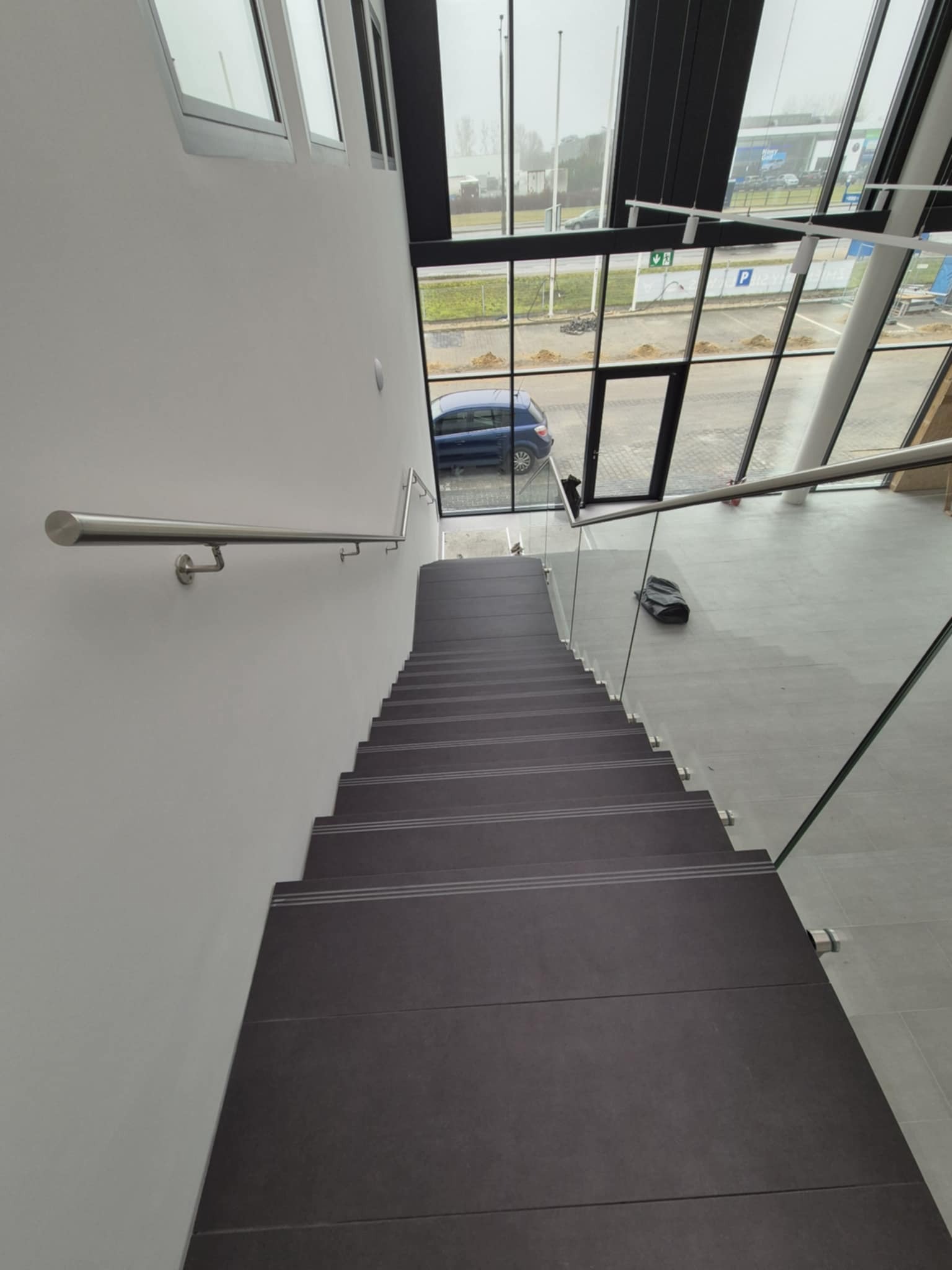Wnętrze biurowca: widok z góry na schody wyłożone ciemnymi płytkami z jasnymi paskami, szklana balustrada, metalowa poręcz, za oknem widoczny parking i budynek z reklamą.