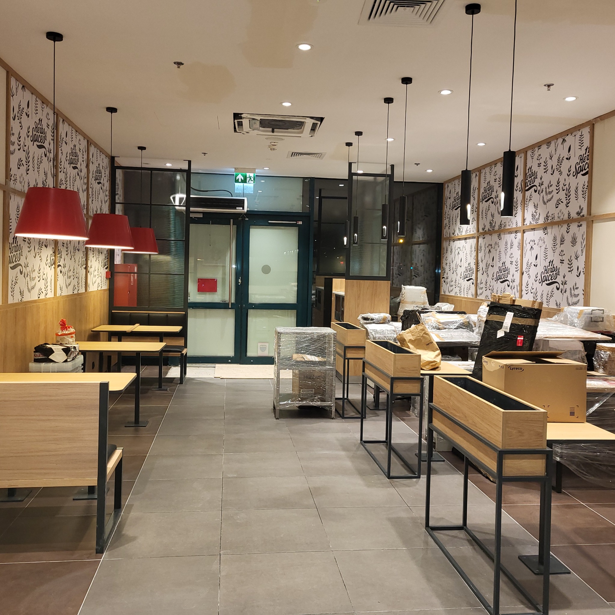 Wnętrze lokalu gastronomicznego w trakcie prac wykończeniowych: meble i dekoracje częściowo zabezpieczone folią, widoczny sufit z kratką wentylacyjną, czerwone lampy wiszące i grafika na ścianach.