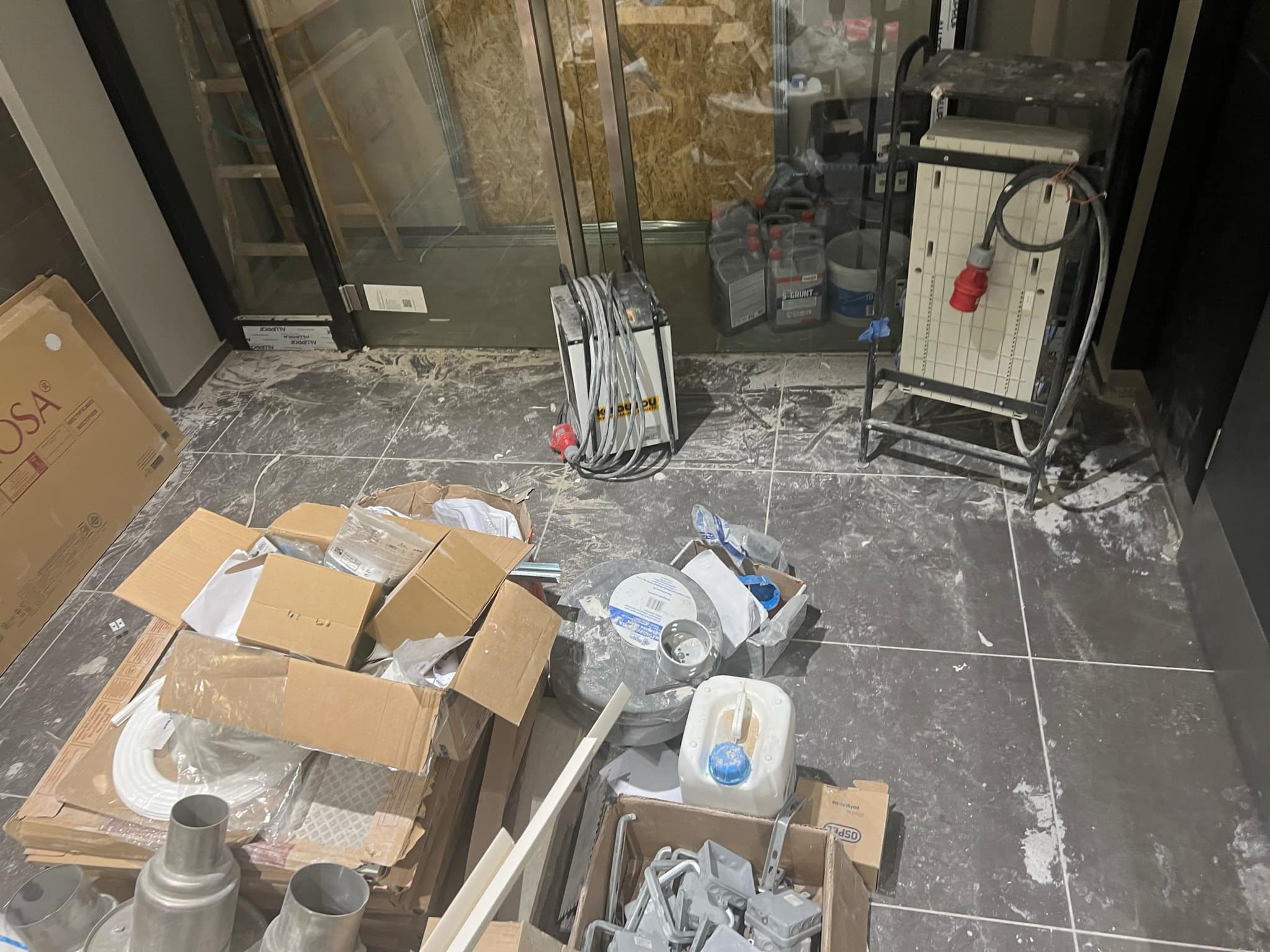 Podłoga w trakcie remontu, widoczne kartony, narzędzia i resztki materiałów budowlanych przy dużym oknie.