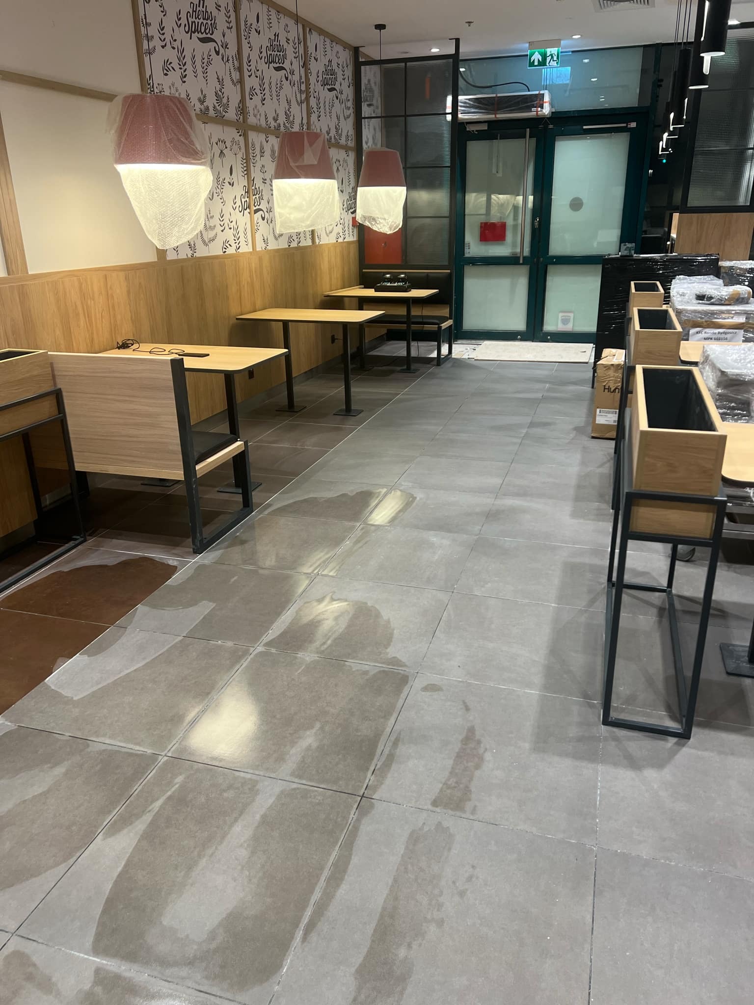 Wnętrze lokalu gastronomicznego w trakcie sprzątania po remoncie: mokra, umyta podłoga z płytek, zabezpieczone lampy, meble częściowo jeszcze ofoliowane, widoczne ślady po pracach budowlanych.