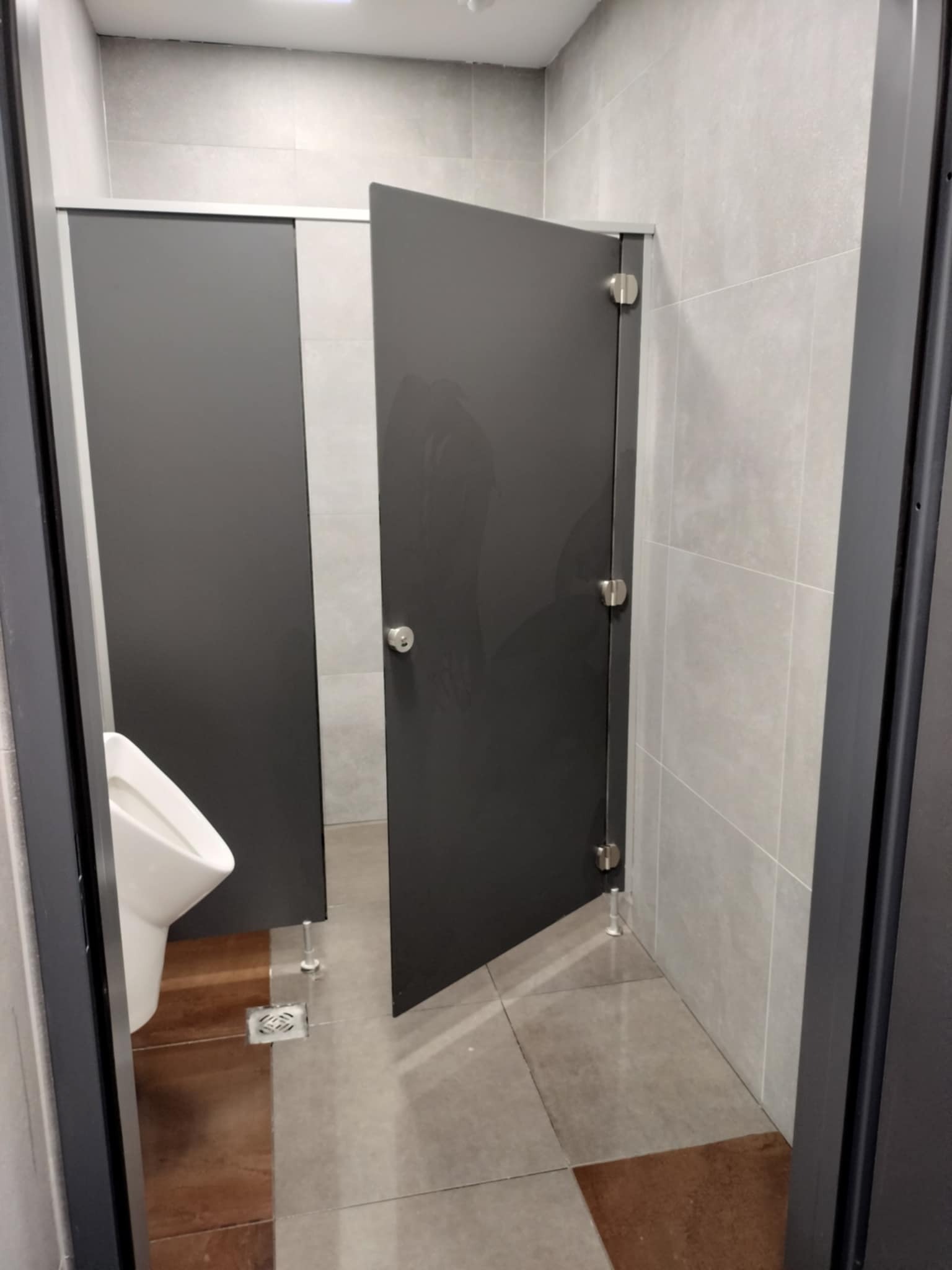 Wnętrze publicznej toalety z kabinami o szarych drzwiach i białym pisuarze, widoczne płytki na ścianach i podłodze.