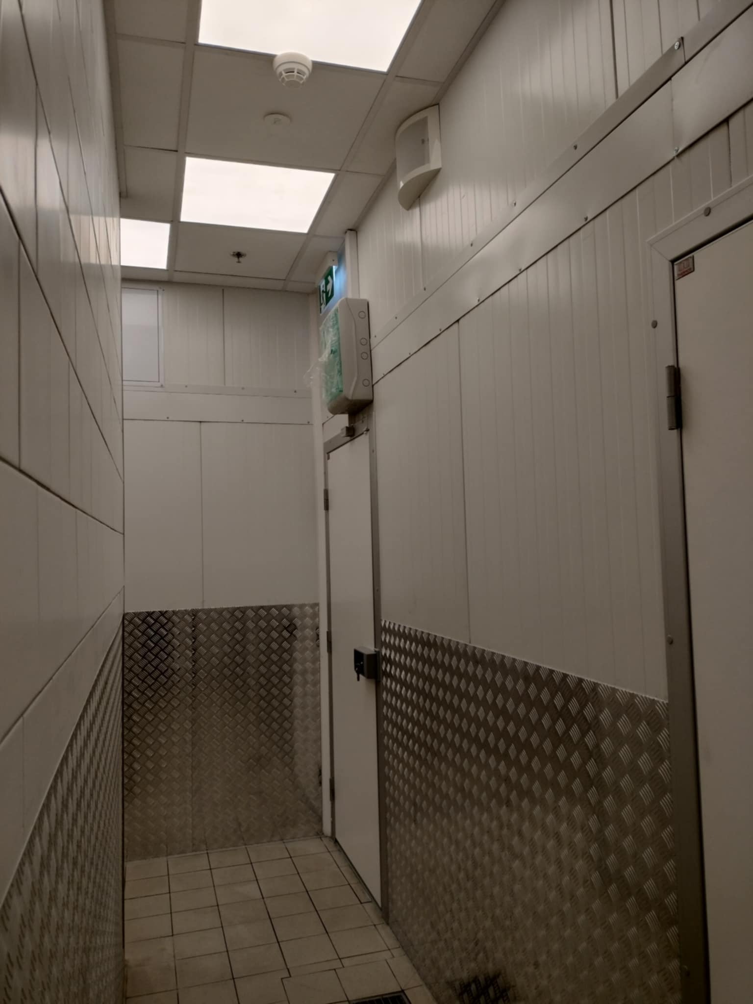 Wnętrze pomieszczenia z panelami ściennymi, widoczne płytki podłogowe i metalowe zabezpieczenie ścian, oświetlenie sufitowe oraz drzwi z zamkiem.
