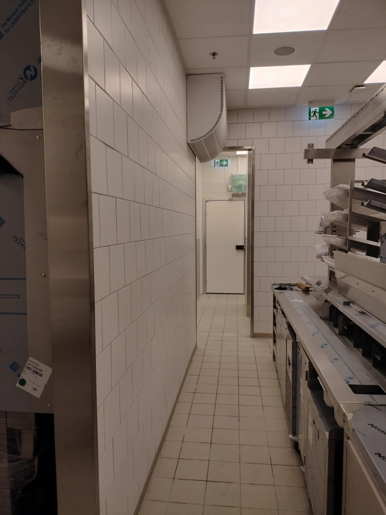 Wnętrze przemysłowe z białymi kafelkami na ścianach i podłodze, widoczne elementy wentylacji i wyposażenia ze stali nierdzewnej, znak ewakuacyjny.