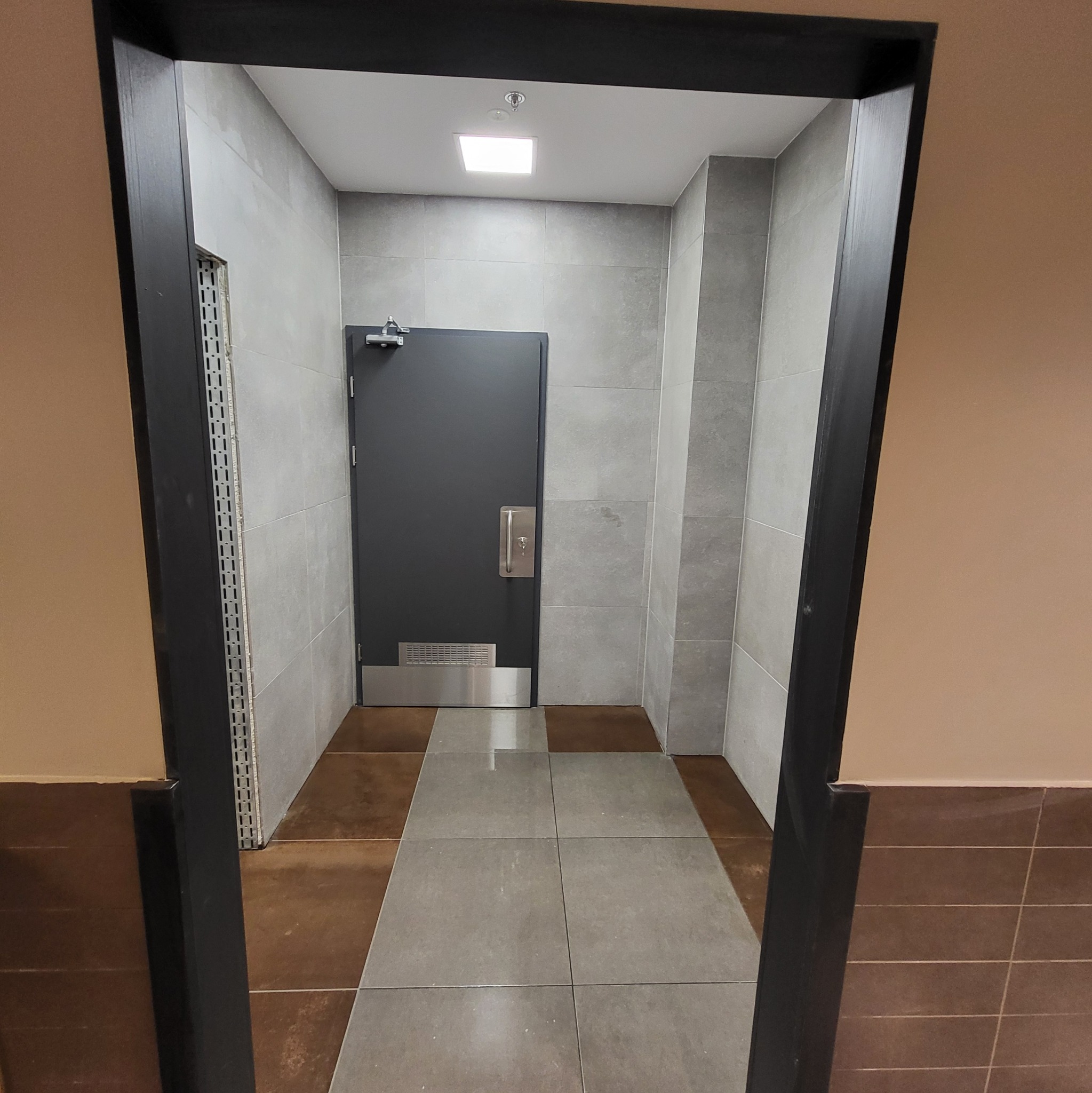 Wnętrze biurowe z widoczną szarą podłogą z płytek, szare ściany z płytek, ciemne drzwi z metalowymi elementami i czarną futryną.