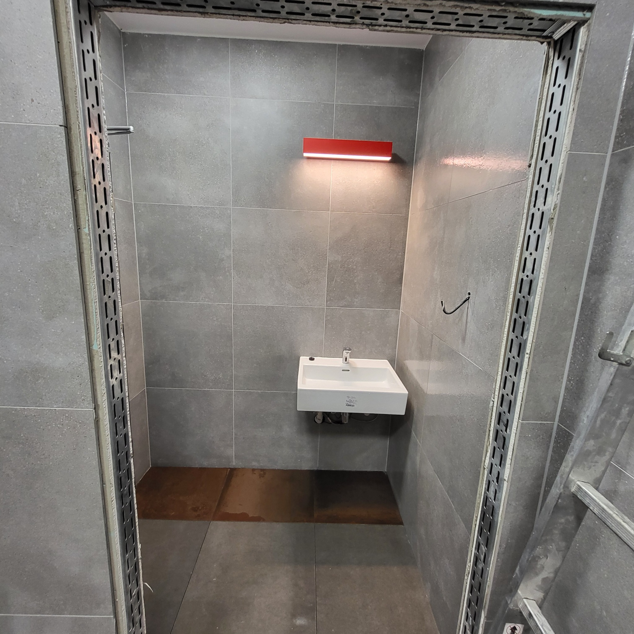 Mała, biała umywalka podświetlona czerwoną lampą w łazience z szarymi płytkami, widoczny metalowy stelaż i mokra podłoga.