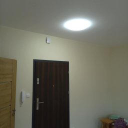 Firma remontowo-budowlane Łukasz Szymczak - Wnętrze pokoju po remoncie: ciemne drzwi wejściowe z metalową ościeżnicą, jasne ściany, drewniana podłoga, biały domofon i szafa w odcieniach drewna.