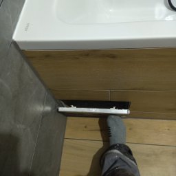 Firma remontowo-budowlane Łukasz Szymczak - Niedokończona instalacja odpływu w szafce pod umywalką KOLO, widoczna noga w skarpecie i spodniach roboczych, płytki imitujące drewno na podłodze i ścianie, szare płytki na ścianie obok umywalki.