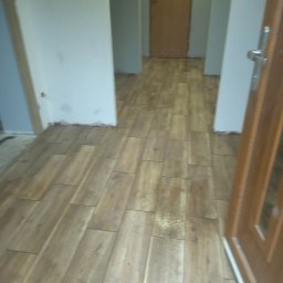 Firma remontowo-budowlane Łukasz Szymczak - Ułożona podłoga z paneli imitujących drewno w korytarzu, widoczna niedokończona listwa przypodłogowa przy ścianie i brązowe drzwi z szybą.