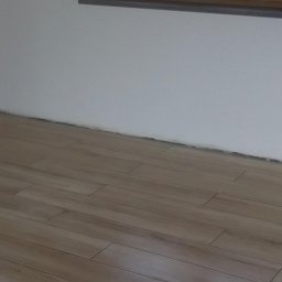 Firma remontowo-budowlane Łukasz Szymczak - Ułożone panele podłogowe imitujące drewno w jasnym, nowym pomieszczeniu z białymi ścianami i oknem w drewnianej ramie.