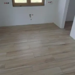 Ułożona podłoga z paneli imitujących drewno w jasnym, pustym pokoju z widokiem na zielone pole za oknem; widoczne gniazdka elektryczne i wyprowadzenia hydrauliczne na ścianie.