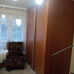 Firma remontowo-budowlane Łukasz Szymczak - Duża, brązowa szafa przesuwna z lustrem w mieszkaniu w Łomowie, obok okna z białymi zasłonami i fotela obitego futrem.