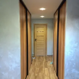 Wąski korytarz z drewnianą podłogą, ścianami pokrytymi dekoracyjną szarą farbą imitującą stiuk oraz zabudową szaf przesuwnych z drewnianymi frontami i czarnymi obramowaniami, zakończony drewnianymi...