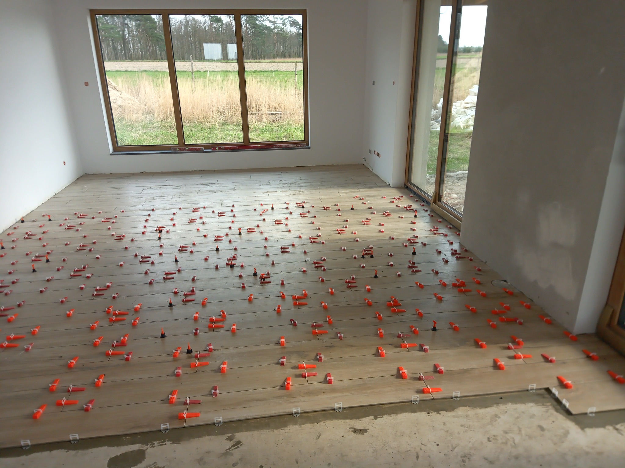 Układanie podłogi z płytek imitujących drewno, z użyciem pomarańczowych klipsów poziomujących, widok na okno z krajobrazem polnym.