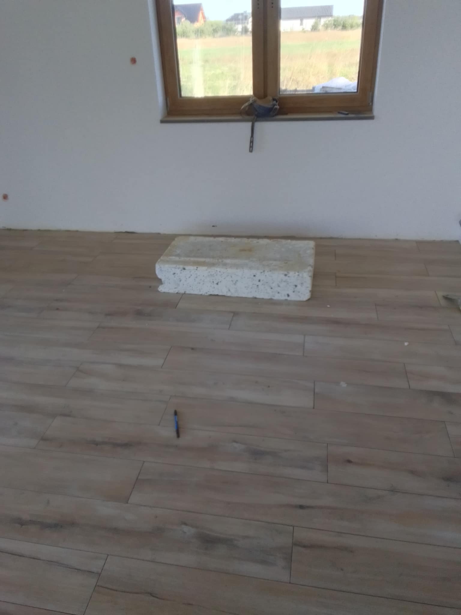 Ułożone panele podłogowe imitujące drewno, widoczny fragment białej ściany, okno z widokiem na pole i niebieski długopis leżący na podłodze.