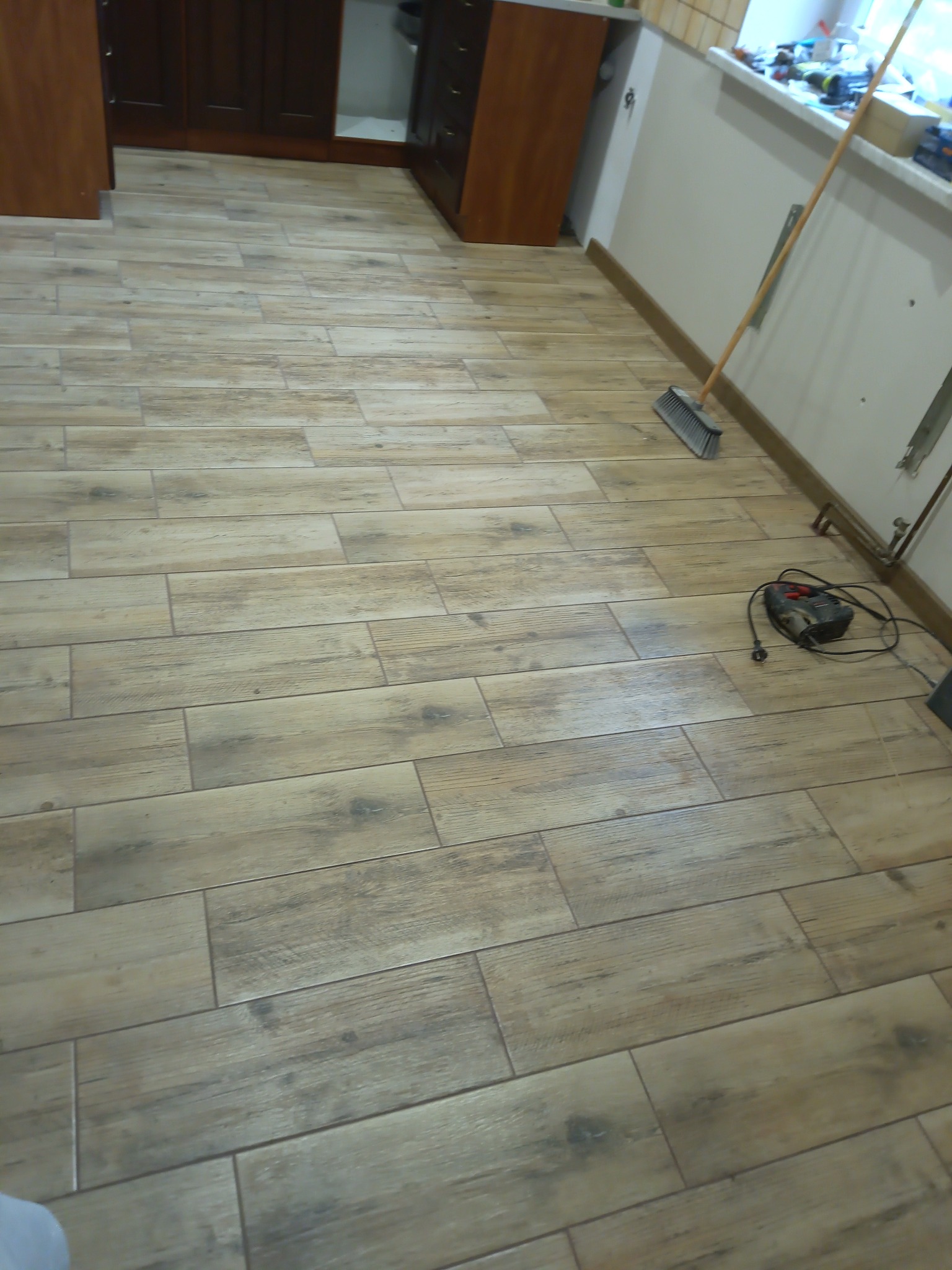 Ułożona podłoga z płytek imitujących drewno, widoczna szczotka i narzędzie do cięcia płytek przy ścianie, fragment szafek kuchennych w tle.