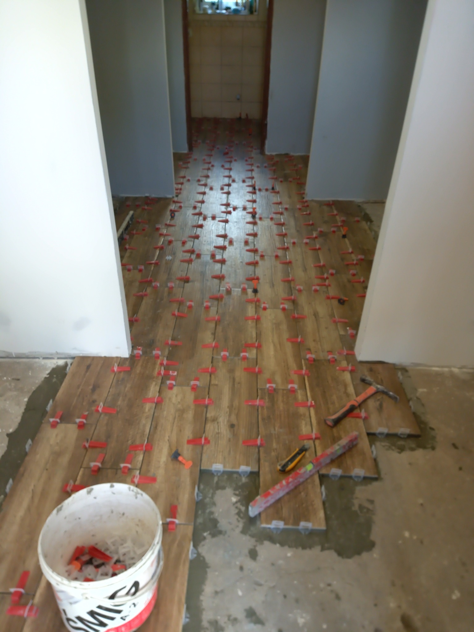 Układanie płytek imitujących drewno z użyciem czerwonych krzyżyków dystansowych, widoczny młotek, poziomica i wiadro z krzyżykami.