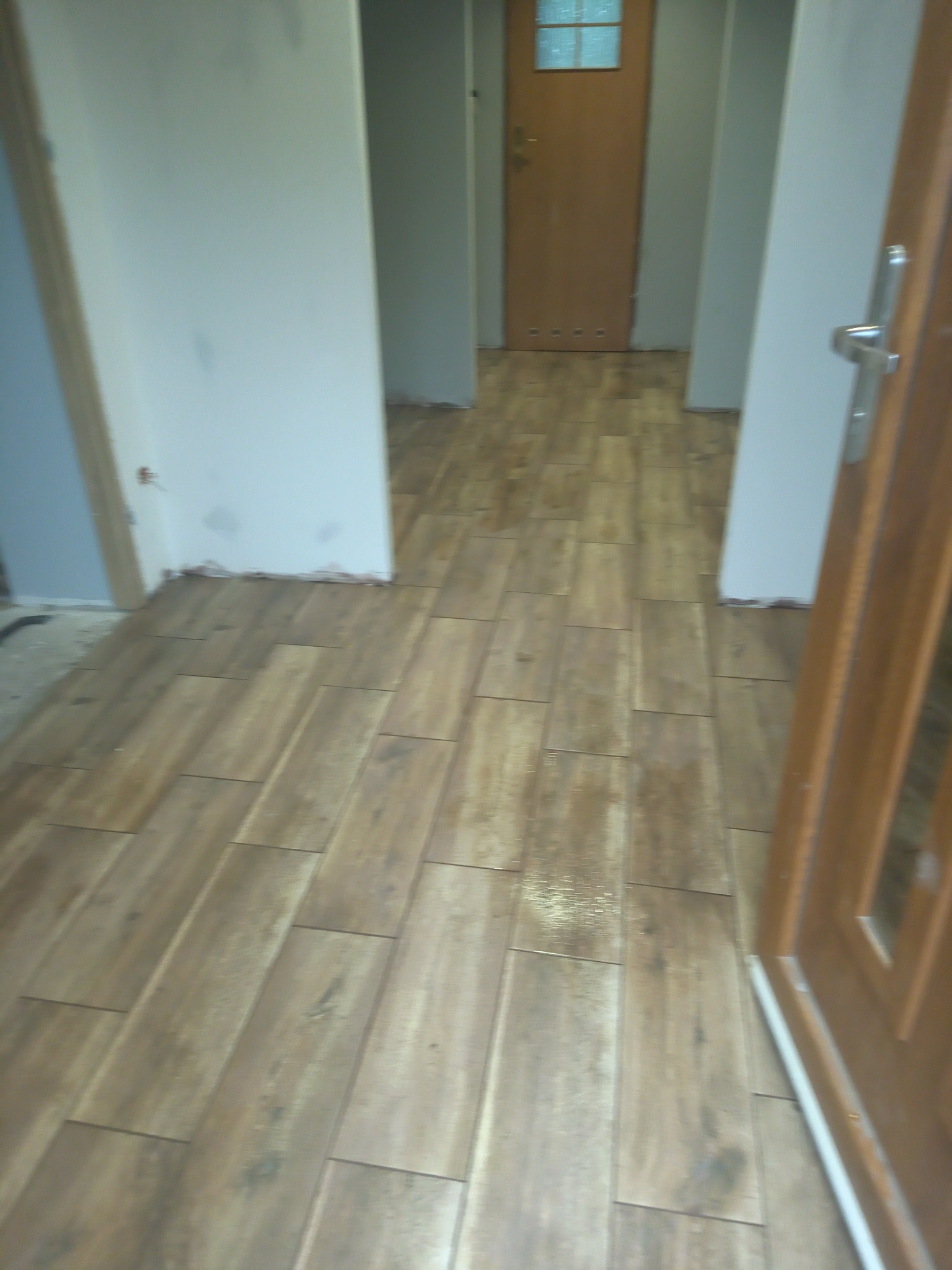 Ułożona podłoga z paneli imitujących drewno w korytarzu, widoczna niedokończona listwa przypodłogowa przy ścianie i brązowe drzwi z szybą.