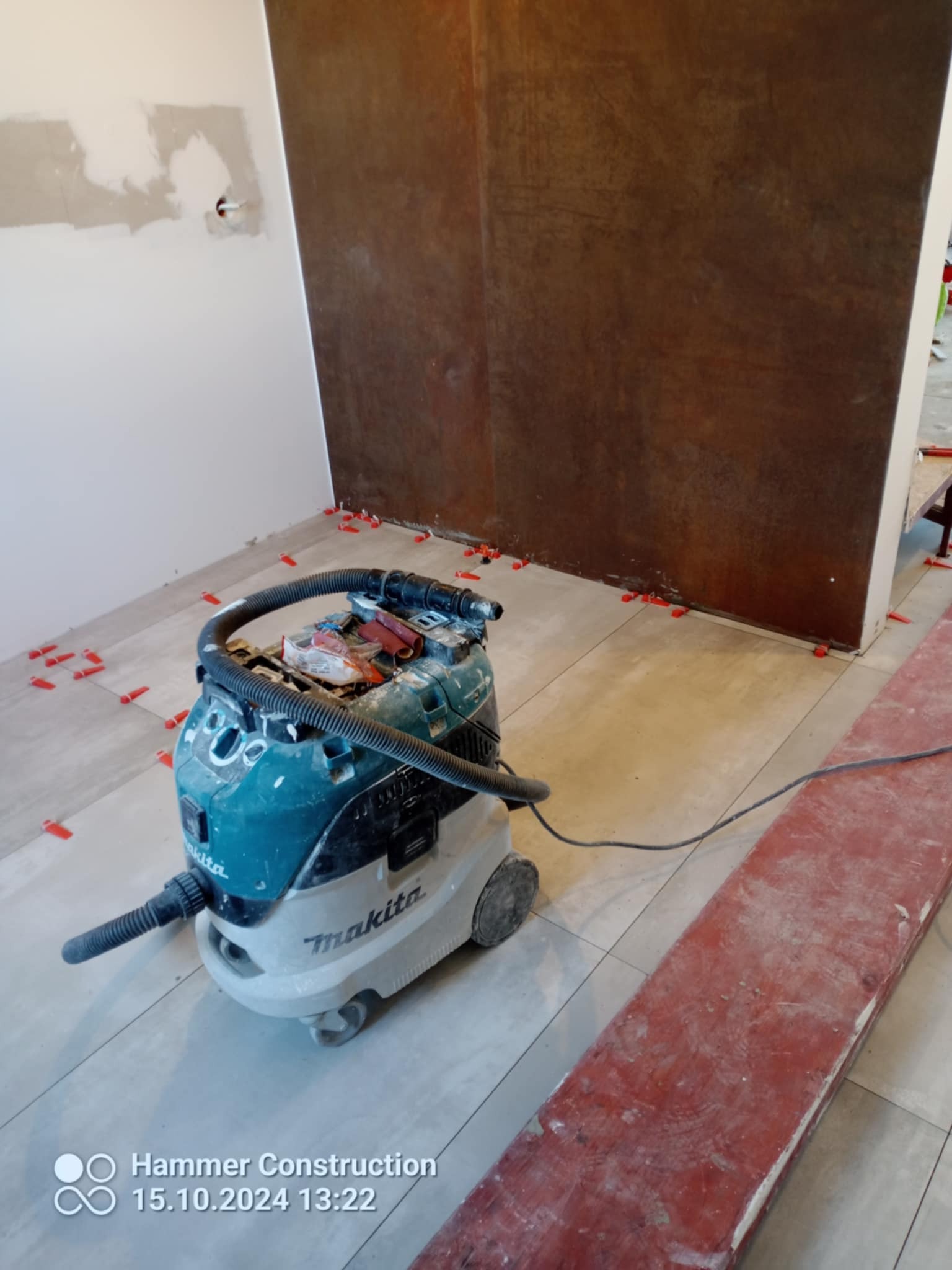 Wnętrze w trakcie układania płytek podłogowych, widoczny odkurzacz budowlany Makita, krzyżyki dystansowe między płytkami i fragment ściany z surową gładzią.