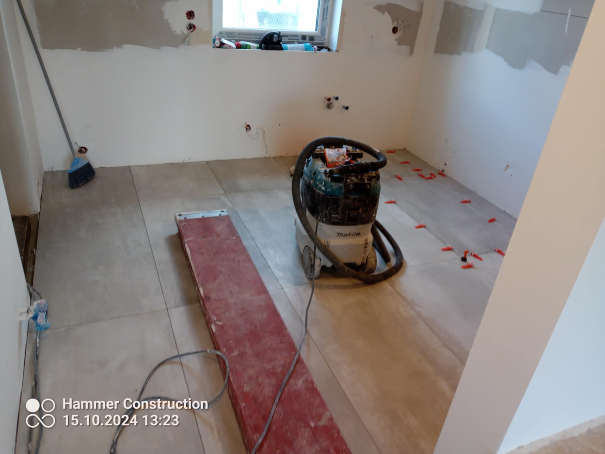 Łazienka w trakcie remontu: ułożone szare płytki podłogowe, widoczny odkurzacz przemysłowy Makita, listwa przypodłogowa oraz czerwone kliny dystansowe do płytek. Ściany częściowo zagruntowane,...