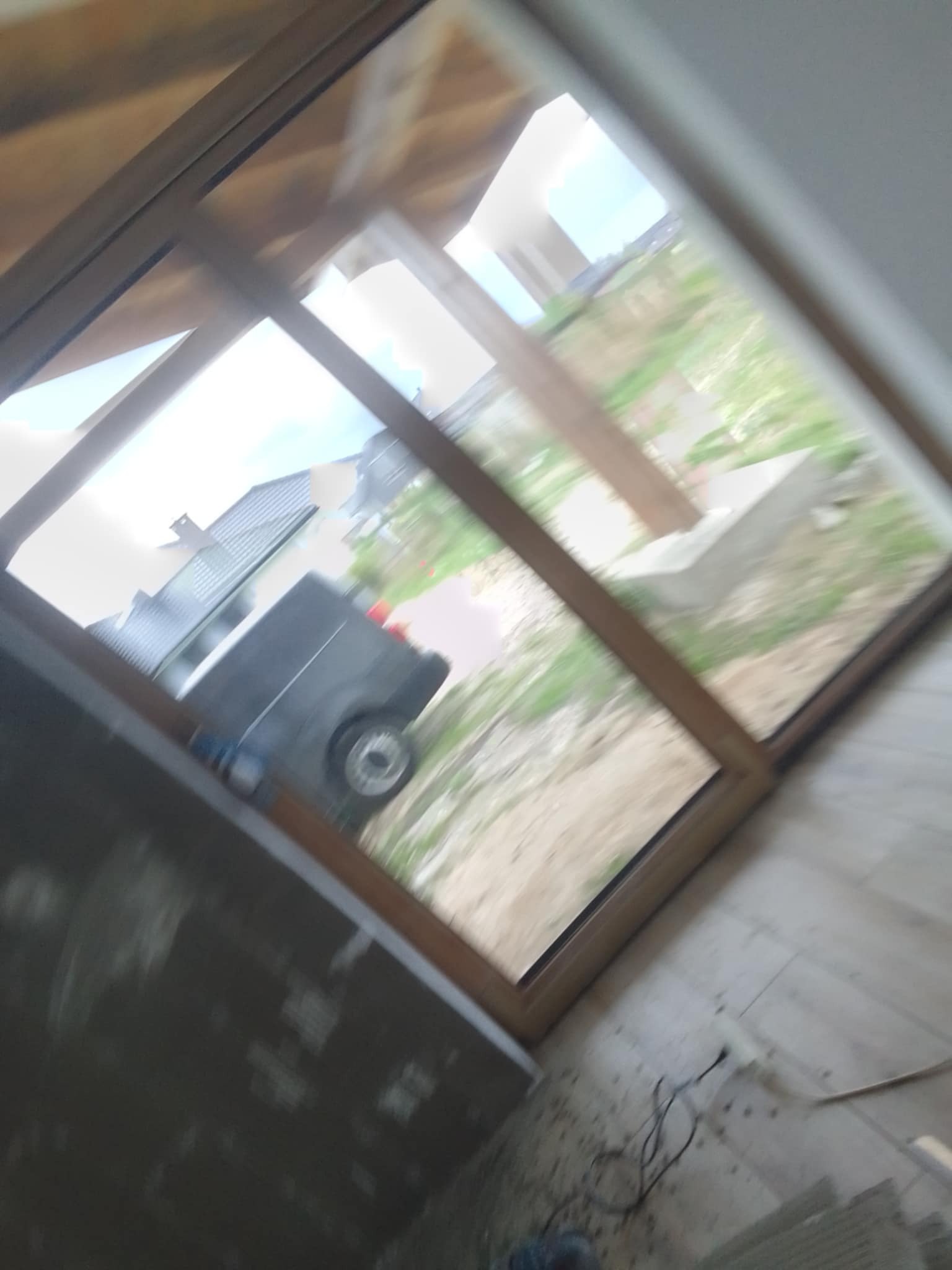 Wnętrze w trakcie prac wykończeniowych: widok przez okno na zewnątrz, fragment podłogi z przewodami, ściana z szarą, surową powierzchnią.  Na zewnątrz widoczny samochód i zieleń.