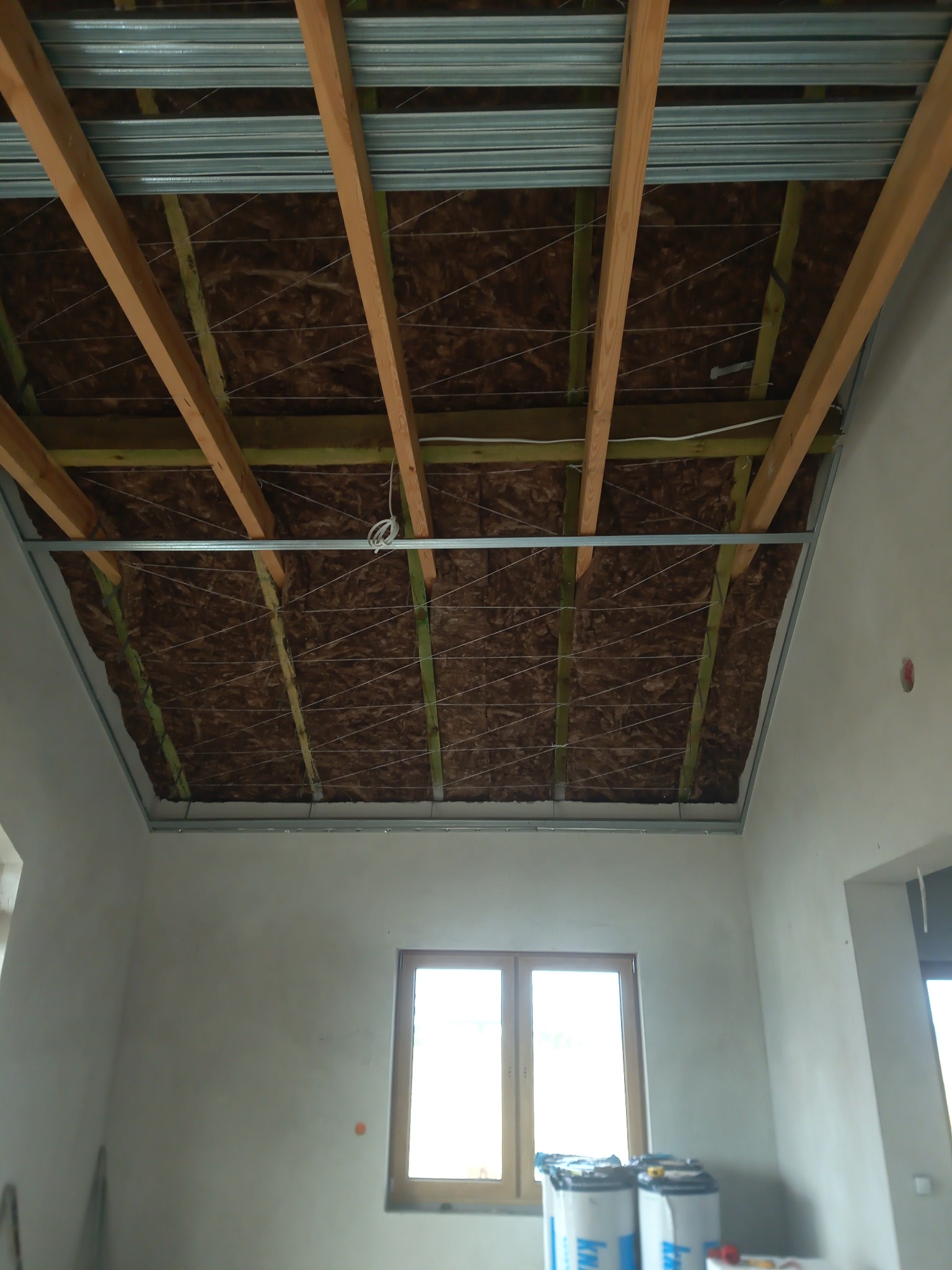 Sufit podwieszany w trakcie montażu: widoczne drewniane belki, izolacja z wełny mineralnej oraz metalowa konstrukcja nośna, w tle okno i worki z materiałami budowlanymi.