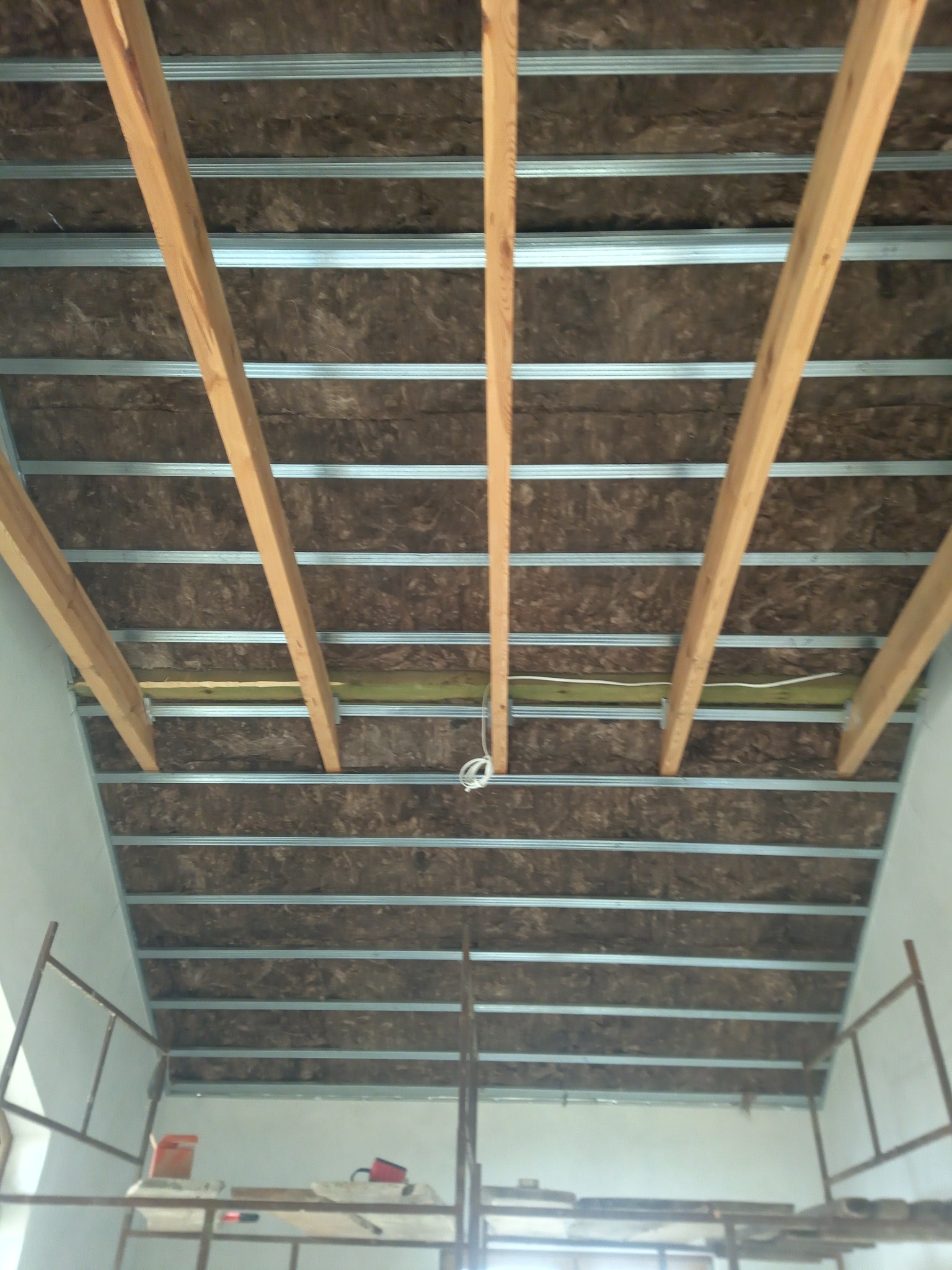 Montaż izolacji z wełny mineralnej i metalowego stelaża pod sufitem podwieszanym na poddaszu, widoczne drewniane belki i fragment rusztowania.