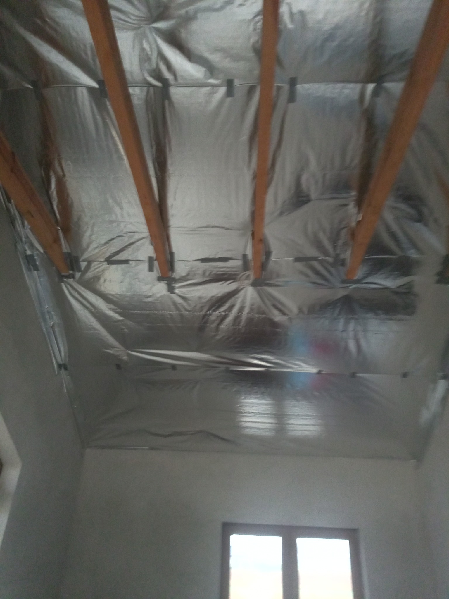 Montaż izolacji termicznej z folii aluminiowej na poddaszu, widoczne drewniane krokwie i okno w ścianie szczytowej.