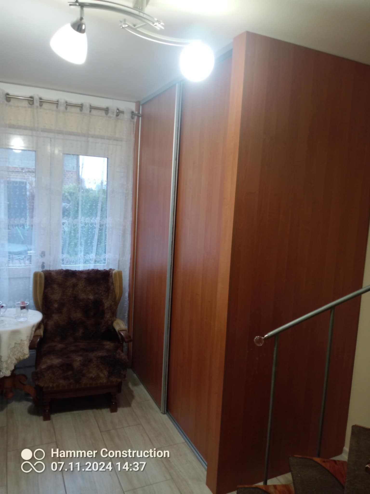 Duża, brązowa szafa przesuwna z lustrem w mieszkaniu w Łomowie, obok okna z białymi zasłonami i fotela obitego futrem.