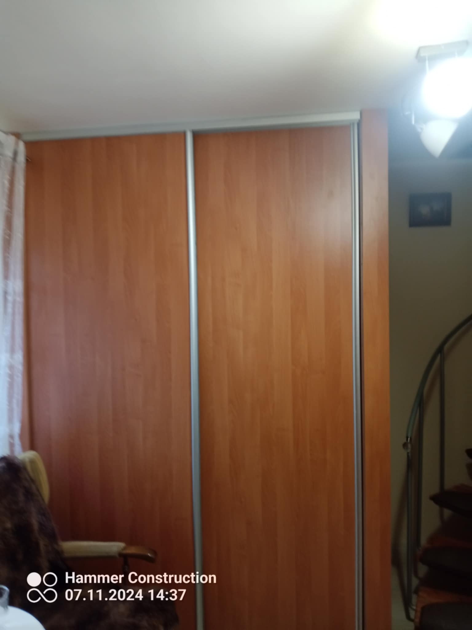 Zamknięta szafa przesuwna z drewnianymi drzwiami w pokoju z krzesłem i schodami, widoczne logo firmy Hammer Construction z datą i godziną.