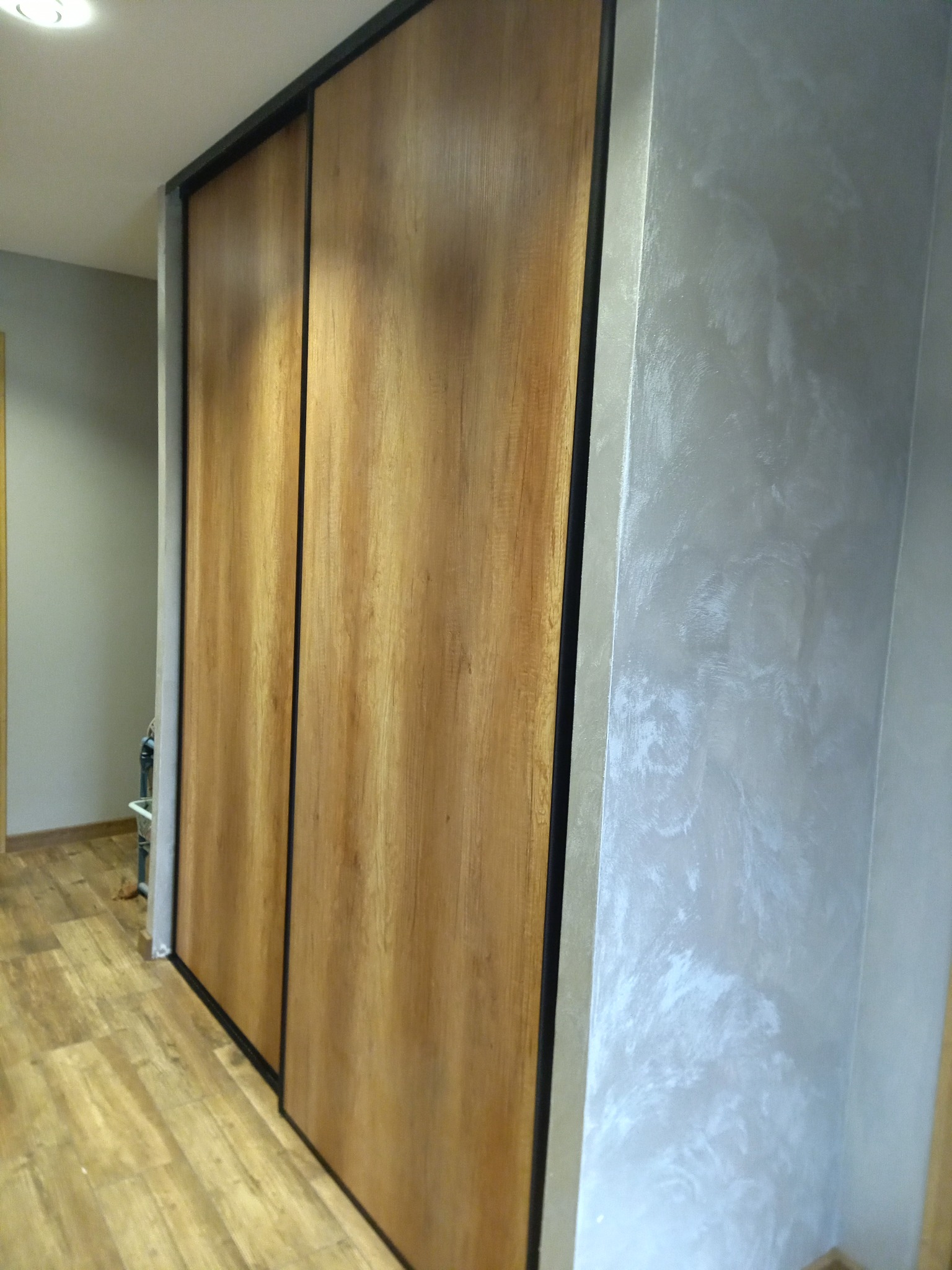 Wbudowana szafa z przesuwnymi drzwiami o drewnianej okleinie, wykończona czarną ramą, na tle ściany pokrytej dekoracyjną szarą farbą strukturalną, dopasowana do drewnianej podłogi.