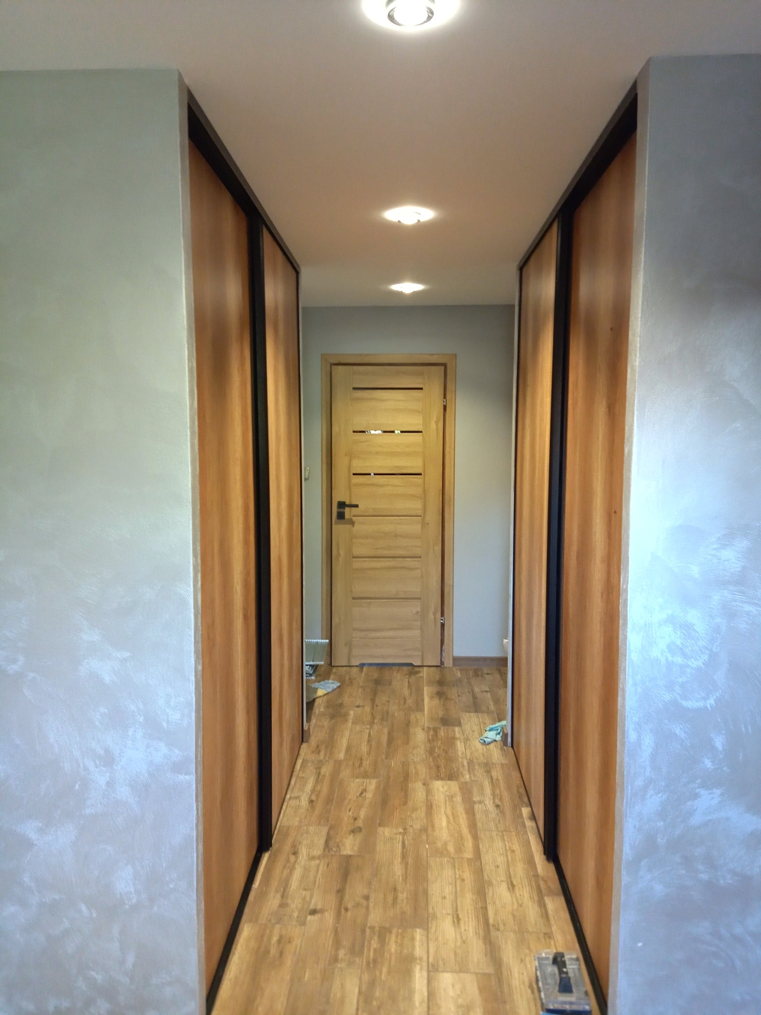 Wąski korytarz z drewnianą podłogą, ścianami pokrytymi dekoracyjną szarą farbą imitującą stiuk oraz zabudową szaf przesuwnych z drewnianymi frontami i czarnymi obramowaniami, zakończony drewnianymi...