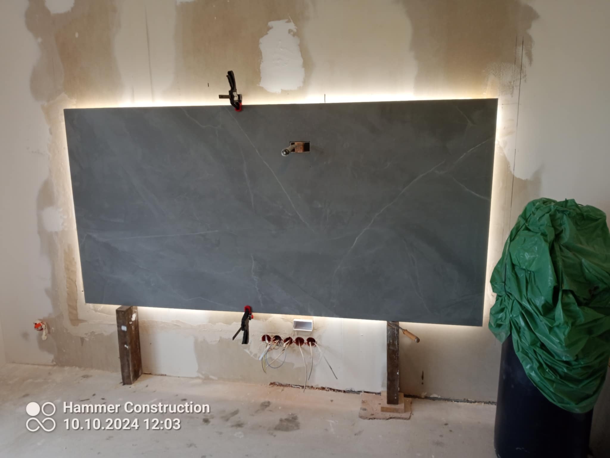 Montaż szarej płyty z marmurowym wzorem, podświetlanej od góry i od dołu, z widocznymi przewodami elektrycznymi i zaciskami, na tle surowej ściany w trakcie prac.