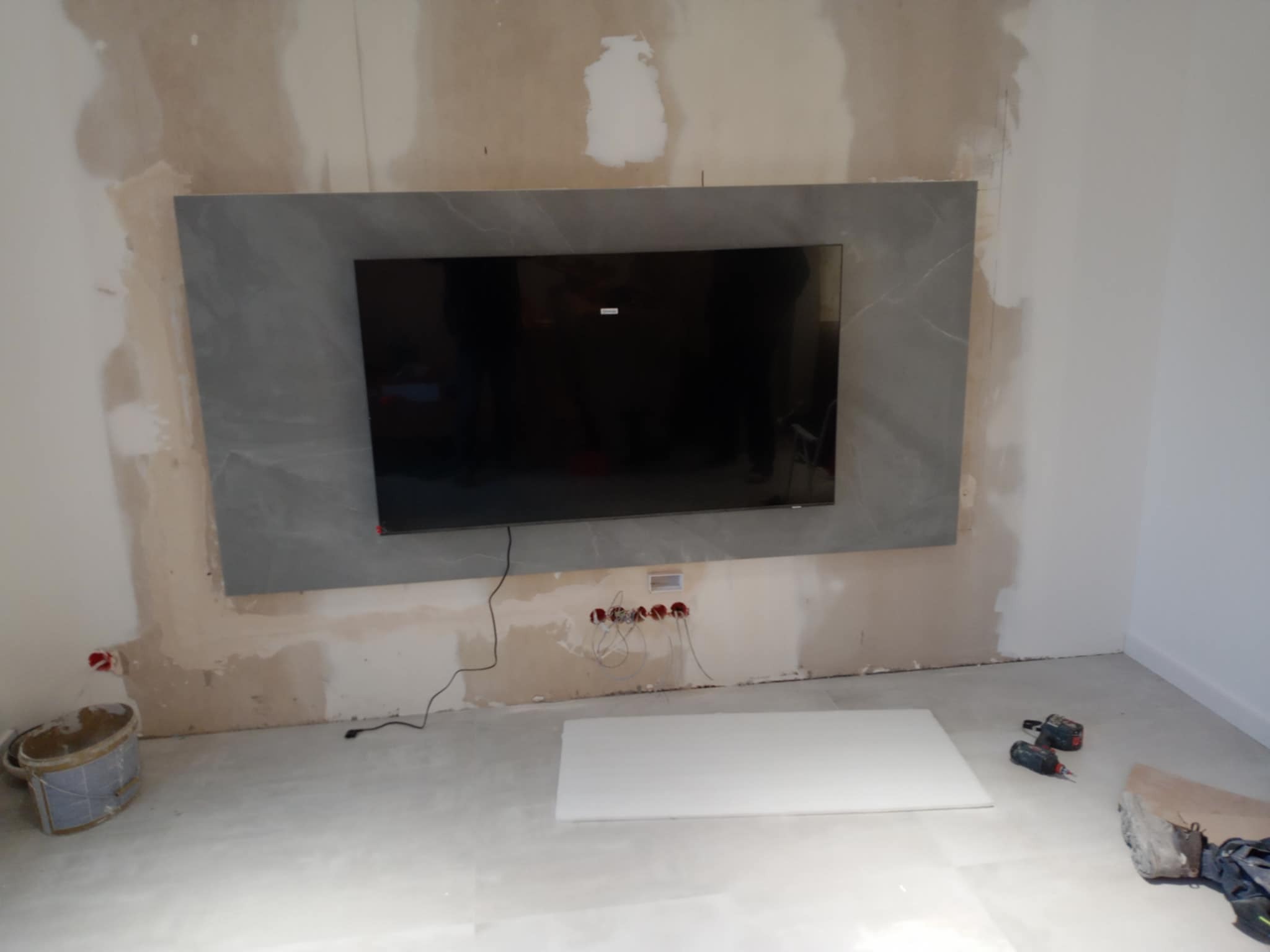 Ściana z zamontowanym telewizorem obłożona szarymi płytami imitującymi kamień, widoczne ślady po gładziowaniu, puszki elektryczne i narzędzia na podłodze, etap prac wykończeniowych.