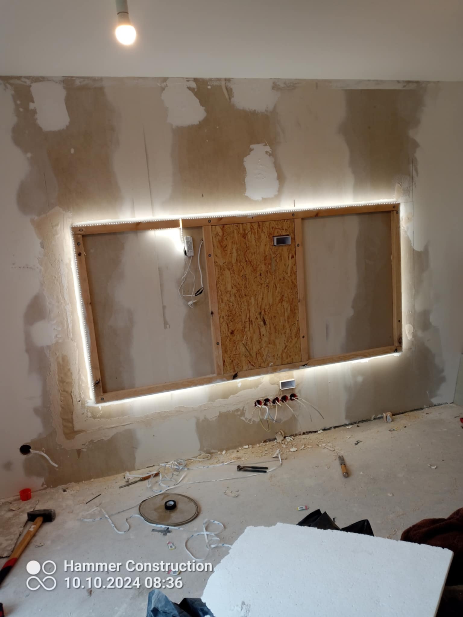 Ściana w trakcie przygotowania do wykończenia z podświetlaną wnęką, widoczne wystające przewody elektryczne i narzędzia na podłodze.