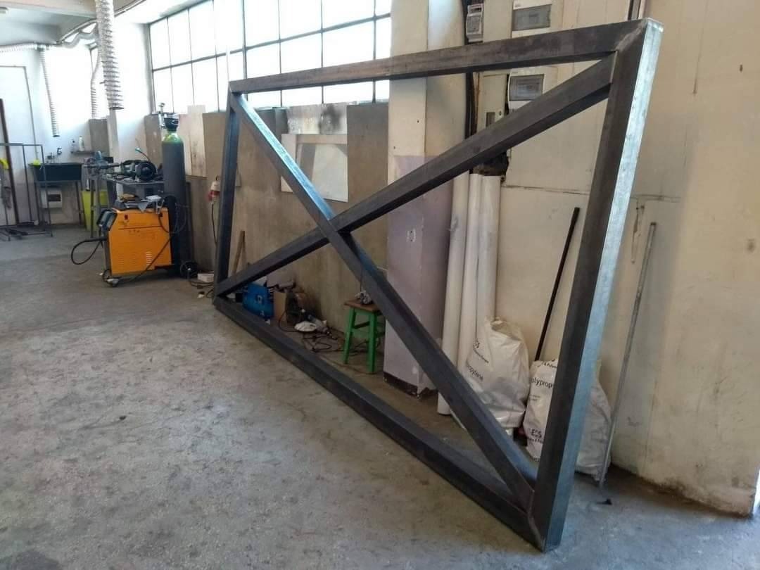 Surowa, spawana konstrukcja metalowa w warsztacie, ukośne wzmocnienia, widoczny sprzęt spawalniczy w tle, worki z materiałami budowlanymi przy ścianie.