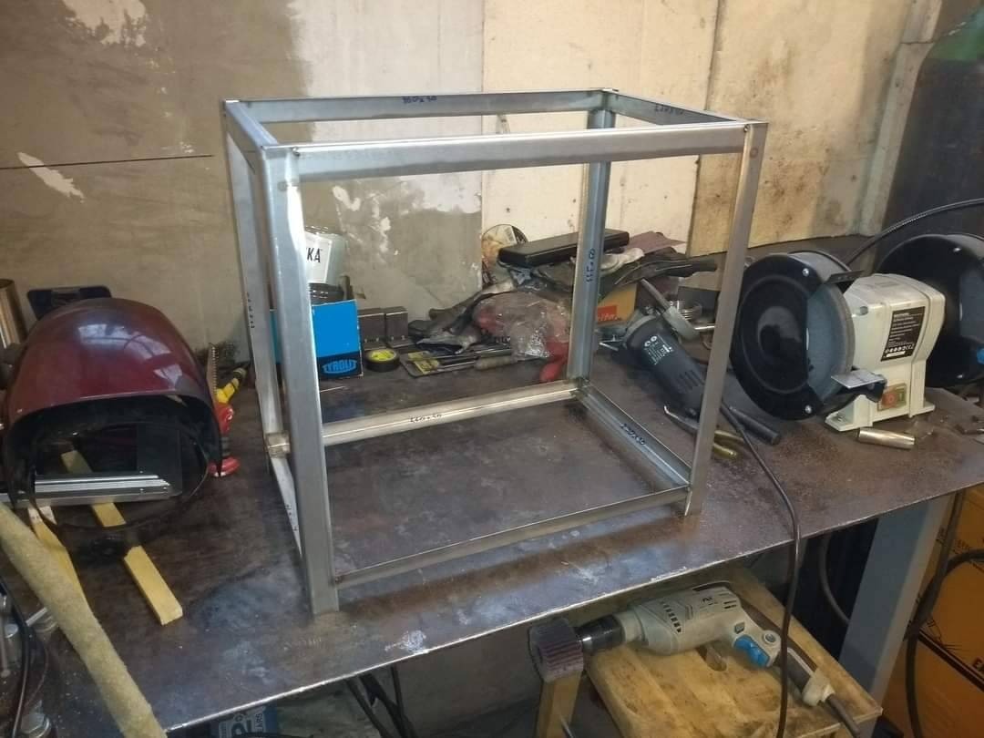 Metalowa rama konstrukcyjna w trakcie budowy na warsztatowym stole, widoczna spawarka, szlifierka i wiertarka, czerwony hełm spawalniczy po lewej.