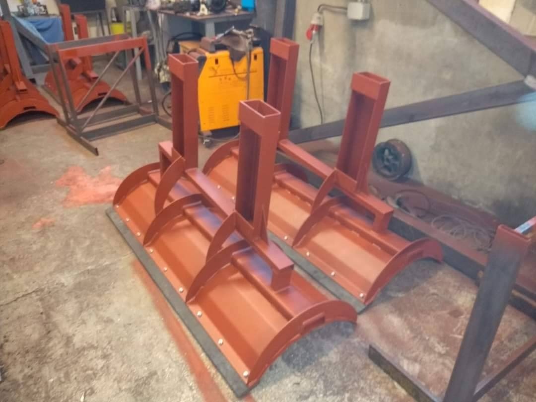Dwa pomalowane na ceglasty kolor, metalowe lemiesze do pługu śnieżnego z przyspawanymi uchwytami, stojące w warsztacie obok spawarki i innych konstrukcji stalowych.