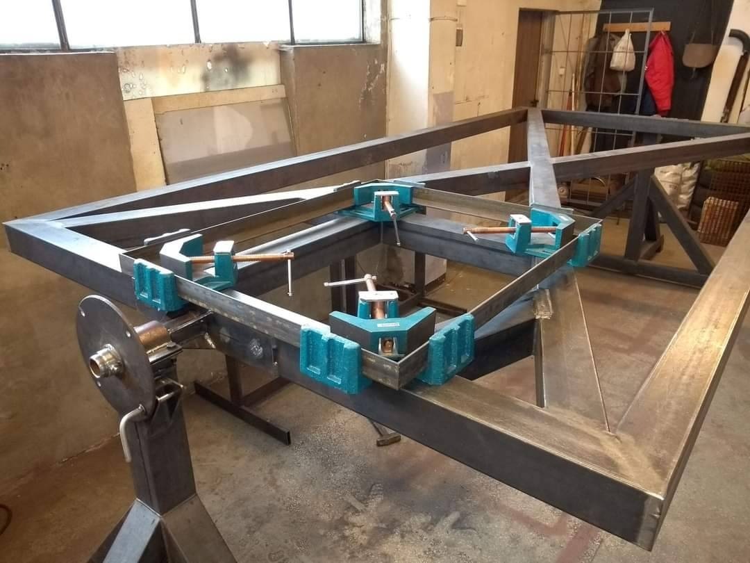 Metalowa rama spawana pod kątem prostym, zabezpieczona niebieskimi zaciskami stolarskimi, ustawiona na metalowym stanowisku spawalniczym w warsztacie.