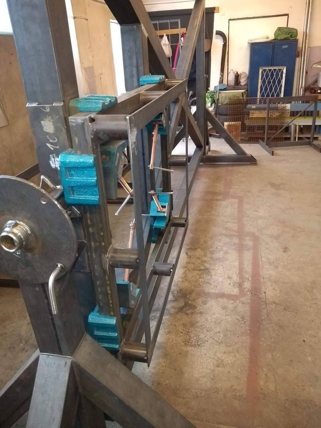 Stalowa konstrukcja spawana, z elementami mocującymi w kolorze turkusowym, w trakcie montażu w warsztacie.