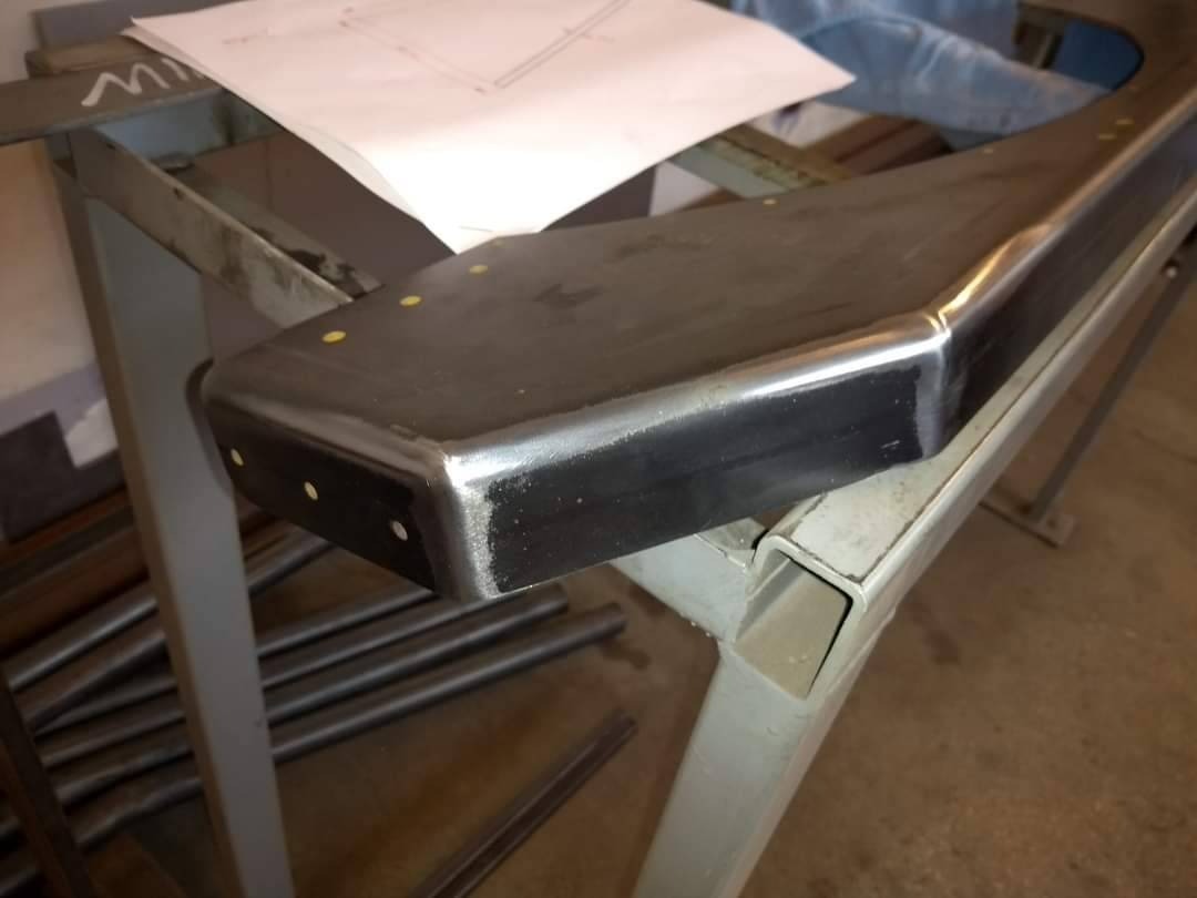 Element metalowy z widocznymi punktami spawania, umieszczony na stole warsztatowym, obok leżą rury metalowe i plan techniczny.