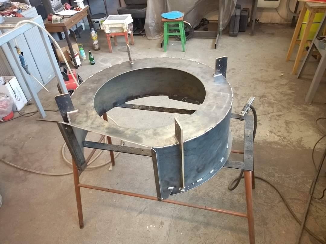 Metalowa konstrukcja w kształcie pierścienia, częściowo spawana, umieszczona na metalowym stojaku w warsztacie.