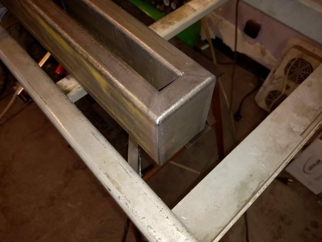 Metalowy profil zamknięty, spawany na rogu, leżący na stole warsztatowym. Widoczne ślady obróbki i niedoskonałości powierzchni.