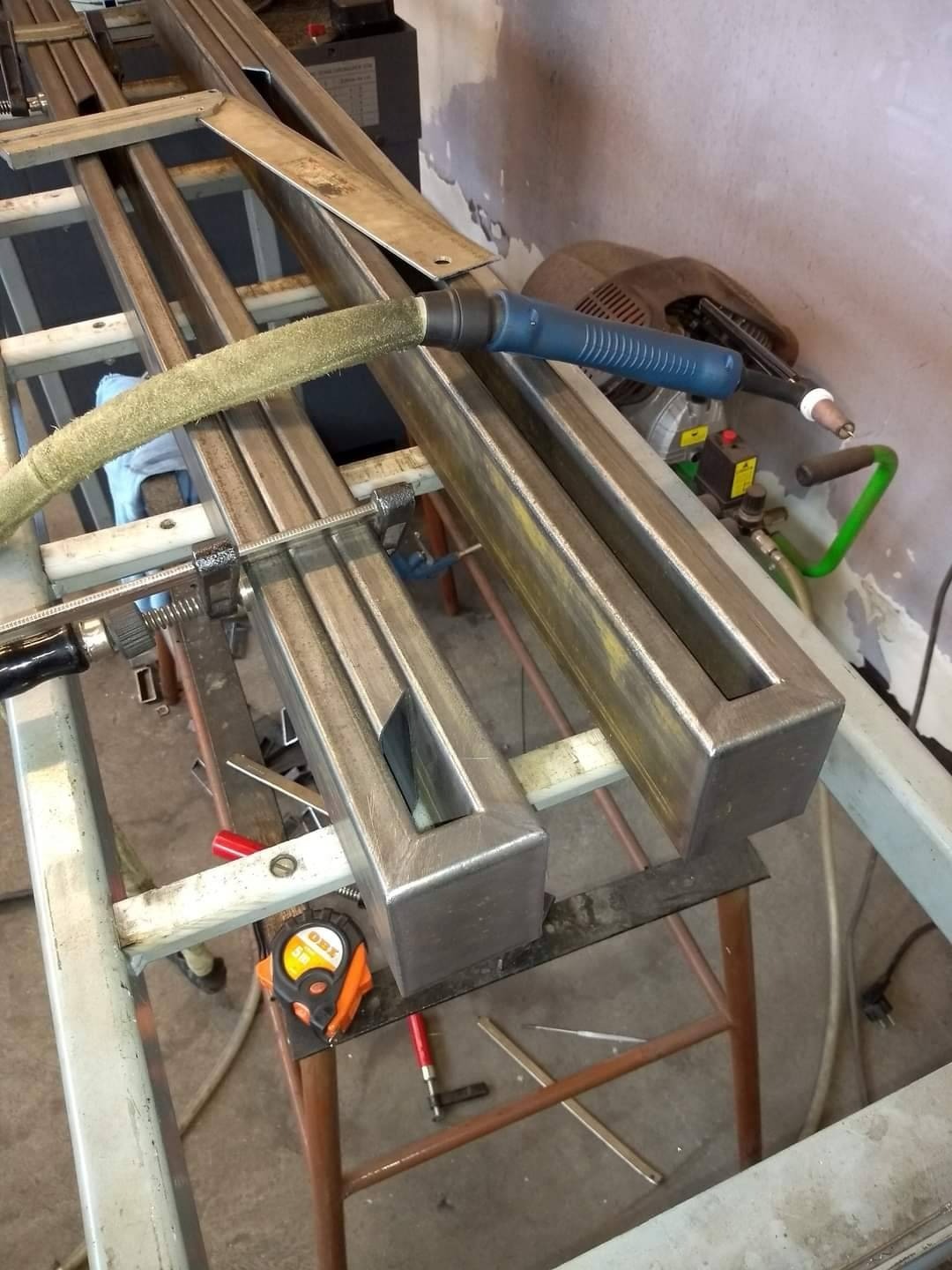 Metalowe profile kwadratowe spawane na stole warsztatowym, widoczny uchwyt spawarki TIG z niebieską rękojeścią i zielonym przewodem, obok leży zwinięta miarka zwijana OBI.