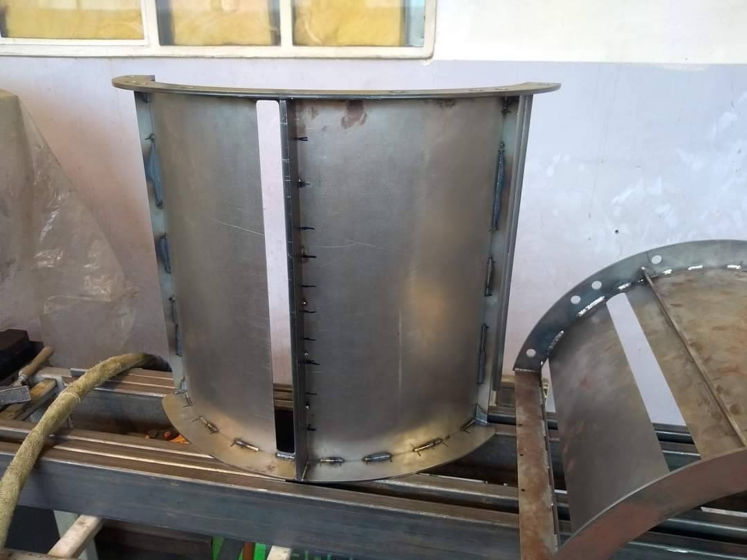 Metalowy element konstrukcyjny o zakrzywionym kształcie, z widocznymi spawami i otworami, leżący na metalowym stelażu w warsztacie.