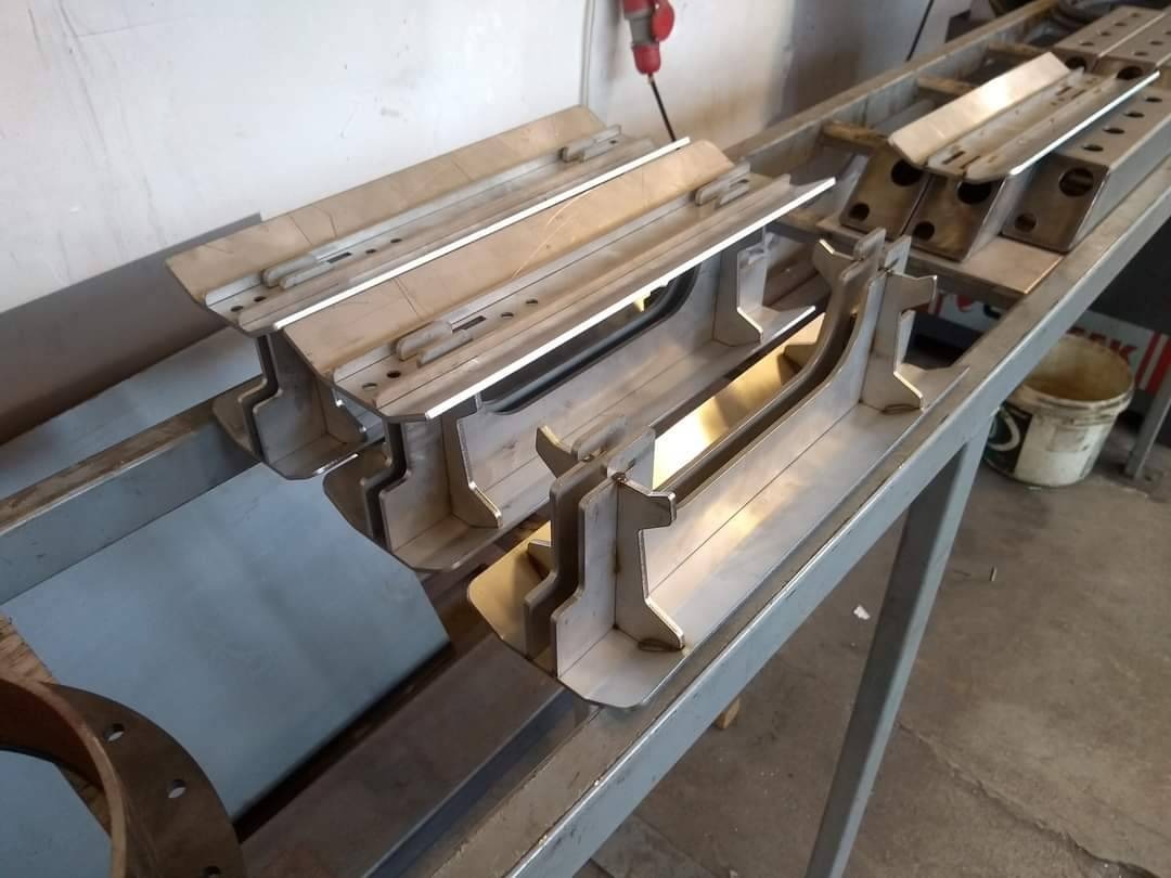 Przygotowane elementy konstrukcji stalowej o nieregularnych kształtach, leżące na metalowym stole warsztatowym, widoczne ślady po obróbce i spawaniu.