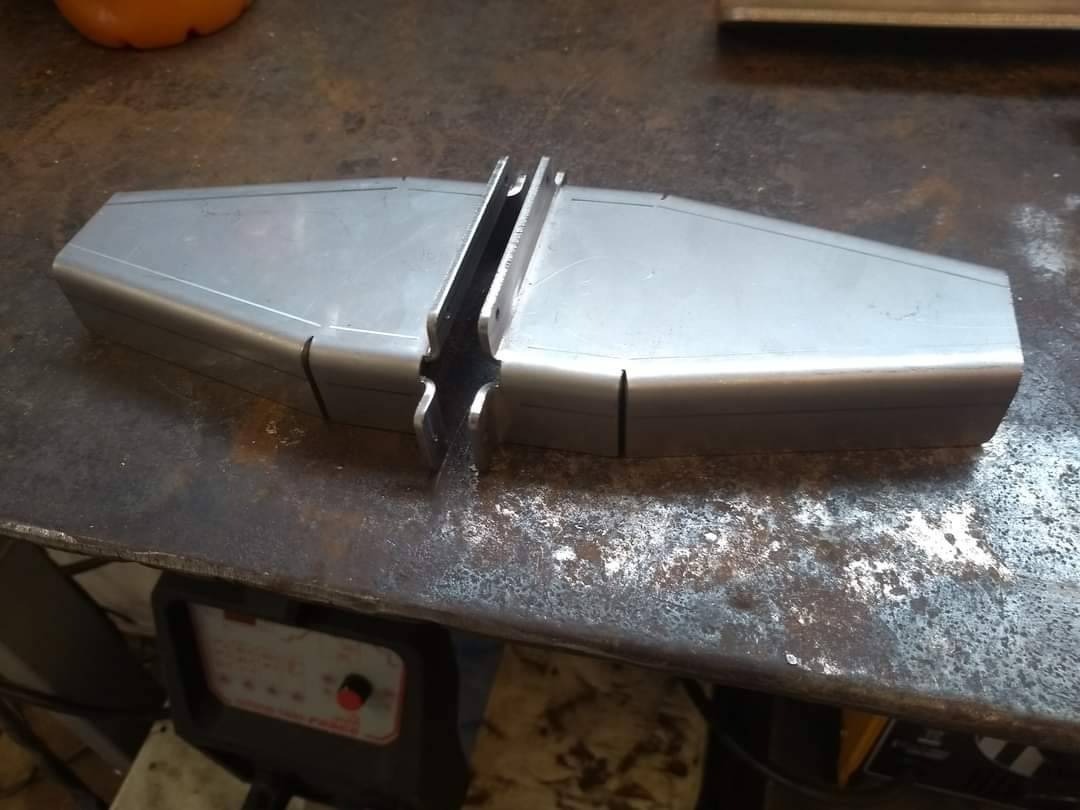 Dwa metalowe elementy z wyciętymi rowkami, przygotowane do spawania na brudnym stole warsztatowym, obok widoczna spawarka.